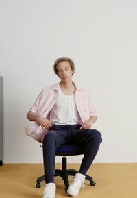 Persona ar svītrainu rozā īso piedurkņu krekls un baltu topu, sēž melnā biroja krēslā, ar tumšiem sporta biksēm un baltām kedām.