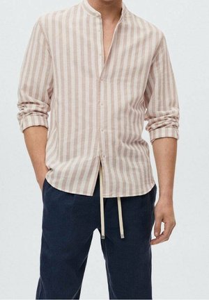 Mężczyzna ubrany w beżowo-białą, pionowo prążkowaną koszulę z długim rękawem, z rękawami podwiniętymi, oraz granatowe spodnie ze ściągaczem, z częściowo wsuniętymi rękami do kieszeni.