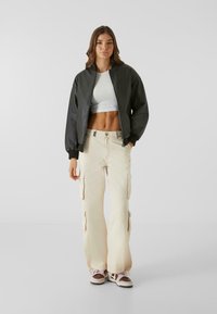 Svart bomberjakke, hvit cropped topp, beige cargo-bukser med lommer, og brune joggesko. Glatte teksturer, avslappet passform og uformelt design.