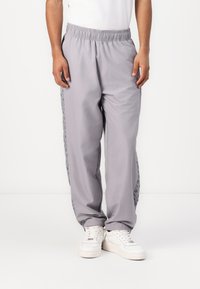 Grijze joggingbroek met een elastische tailleband, voorzien van zijkanten in een gemusterd stof en zijzakken. Gekleed met witte sneakers.
