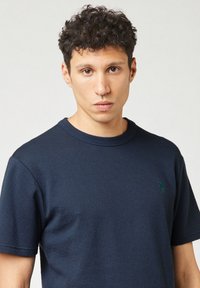 T-shirt a maniche corte blu navy realizzato in tessuto testurizzato, con un piccolo logo di un albero verde sul lato sinistro del petto. Collo rotondo e vestibilità rilassata.