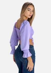 Fliederfarbenes, kurz geschnittenes Oberteil mit Puffärmeln, geraffter Vorderseite und offenem Rückendetail. Kombiniert mit hoch taillierten Jeans aus Denim. Glatte Textur.