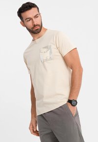 Camiseta de algodón beige con un bolsillo a parche de camuflaje. Mangas cortas, diseño de cuello redondo, combinada con pantalones grises y un reloj negro.