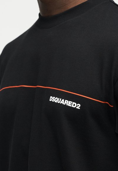 Μαύρο μπλουζάκι με στρογγυλή λαιμόκοψη, λεπτή οριζόντια πορτοκαλί γραμμή και λευκό λογότυπο "DSQUARED2" στο στήθος, φορεμένο από ένα άτομο.