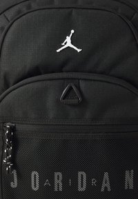 Svart ryggsäck med texturerat tyg, nätficka, vit Jumpman-logotyp och kontrasterande dragkedjeförslutning med svartvitt mönster.