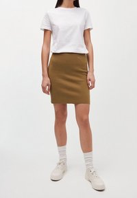 Gonna aderente verde-marrone realizzata in tessuto testurizzato, abbinata a una maglietta di cotone bianca e scarpe da ginnastica bianche, indossata con calzini bianchi.