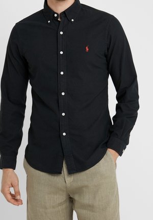 Homme portant une chemise noire à manches longues boutonnée avec un logo rouge sur la poitrine et un pantalon beige, une main dans la poche.