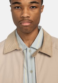 Chaqueta beige con un cuello de pana sobre una camisa a rayas azul y blanca. La camisa presenta una tapeta de botones con botones a juego.