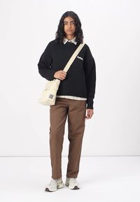 Zwart gebreid trui met geribbelde zoom, beige crossbody tas, bruine broek en witte sneakers. Subtiele merklogo op de trui.