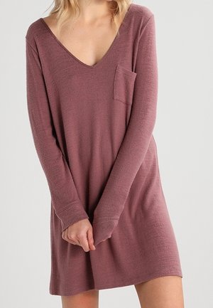 Kobieta ubrana w różową sukienkę o długości do kolan z długimi rękawami, z dekoltem w serek i małą kieszonką na piersi, stojąca na jednolitym tle.