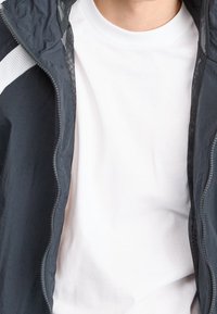 Tummanharmaa kevyt takki verkkovuorillisella hupulla, jossa valkoinen korostusraita, päällä yksivärinen valkoinen t-paita. Takissa näkyvä vetoketju.