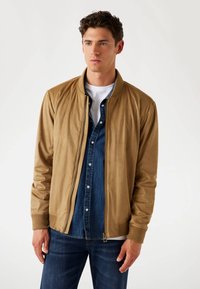 Tan bomberjack met ribgebreide manchetten en kraag, met een ritssluiting aan de voorkant, gecombineerd met een denimbloes eronder, gedragen met donkere jeans.