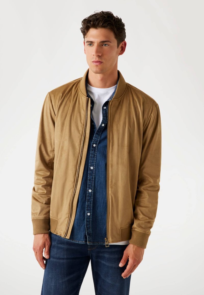 Tan bomberjack met ribgebreide manchetten en kraag, met een ritssluiting aan de voorkant, gecombineerd met een denimbloes eronder, gedragen met donkere jeans.