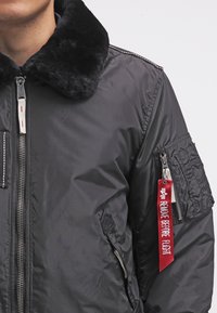 Bomberjacka i svart nylon med krage i fuskpäls, dragkedja och en ficka med patch på ärmen med en röd etikett märkt "TA BORT INNAN FLYGNING."