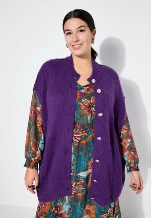 Lilla strikket cardigan med ribbet krave, frontlukning med knapper og struktureret stof, båret over en farverig blomstermønstret kjole.