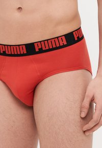 Piros pamut alsó nadrág, fekete derékpánttal, amelyen a "PUMA" logó piros színben található. Az anyag sima tapintású, testhezálló formájú, lapos varrásokkal.