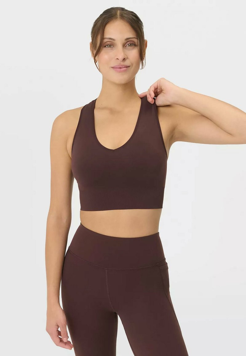 Jeune femme portant un haut court sans manches marron foncé et des leggings taille haute, souriante avec une main touchant son épaule.