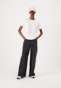 Lässiges Outfit mit einem cremefarbenen T-Shirt mit pinkem Aufdruck, weiten schwarzen Hosen und einer pinken Kappe mit weißem Logo. Sneakers runden den Look ab.