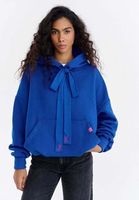 Hanorac albastru cu glugă, buzunar frontal, detaliu de fundiță la gât și logo de brand roz. Material moale, croială lejeră și mâneci lungi.