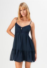 Jonge vrouw met blond haar, gekleed in een mouwloze marineblauwe jurk met kantdetails en een strik op de borst, staande tegen een effen witte achtergrond.