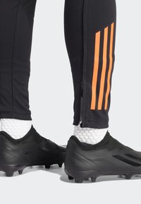 Un pantalon de sport noir avec un design à trois rayures oranges et des fermetures éclair aux chevilles, associé à des chaussures de sport noires et des chaussettes blanches texturées.