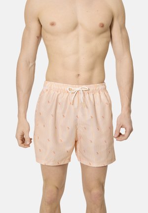 Hombre con pantalones cortos de baño a rayas en naranja claro y blanco con pequeños estampados de flamencos y cintura con cordón blanco, de pie frente a un fondo liso.