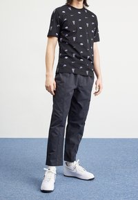 Osoba oblečená v černém tričku Nike s logy a textem, tmavých kalhotách a bílých teniskách Nike, stojící na modré kobercové podlaze proti bílé zdi.