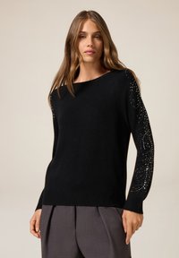 Maglione nero realizzato in morbido tessuto a maglia, con maniche lunghe dotate di dettagli decorativi e orlo a costine. Design semplice e pulito.