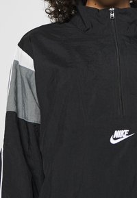 Svart Nike vindjacka med halvdragkedja framtill, med grå och vita paneler, hög krage och en liten vit logotyp på bröstet.