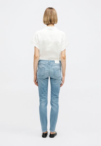 Personne aux cheveux blonds courts vue de dos, portant une chemise blanche à manches courtes, un jean skinny bleu clair et des chaussures noires