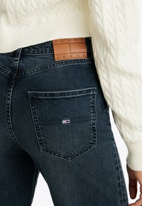 Vaqueros azul oscuro con una parche de cuero en la cintura etiquetado como "TOMMY JEANS." Cuenta con un bolsillo trasero que tiene un pequeño logotipo rojo, blanco y azul.