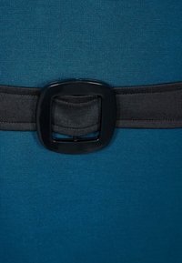 Tyrkysová látka se hladkým povrchem obsahuje černý elastický pás zabezpečený čtvercovou sponou uprostřed.