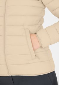 Beige pufferjakke med horisontalt quiltet mønster, lynlås lukning og sidelomme. Har fitted manchetter og en blød, glat tekstur.