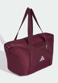 Μπορντό αθλητική τσάντα duffel με διπλές λαβές, πλαϊνή τσέπη με φερμουάρ, ρυθμιζόμενο ιμάντα και μικρό λευκό λογότυπο στο μπροστινό μέρος.
