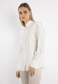 Camicia bianca a maniche lunghe con bottoni, realizzata in tessuto leggero, con colletto classico e dettagli di pieghe sottili sulle spalle.