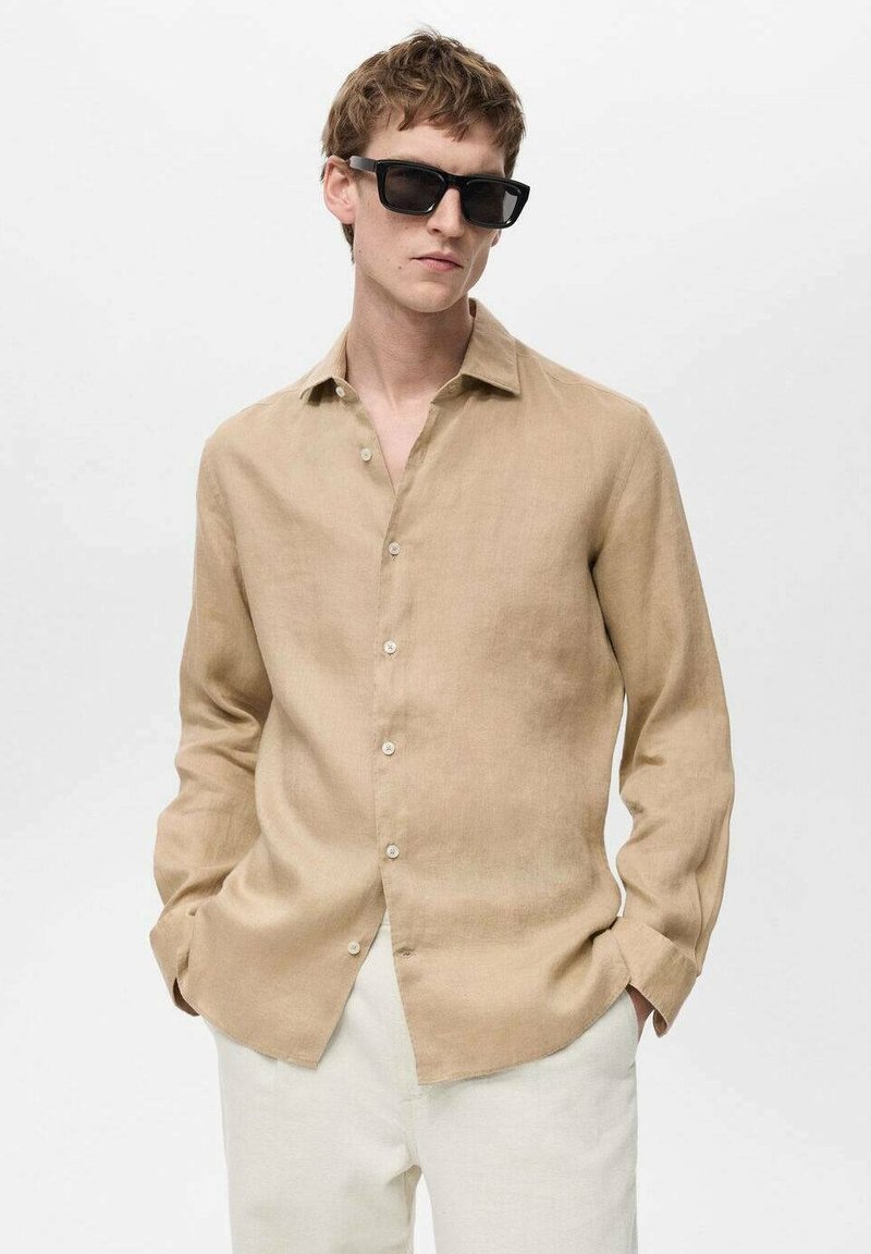 Camicia in lino beige con colletto classico, maniche lunghe e chiusura a bottoni. Texture liscia con vestibilità comoda. Indossata con pantaloni bianchi.