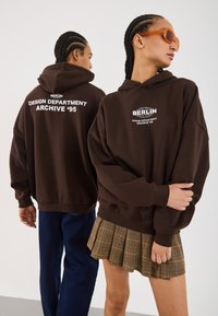 Bruine hoodie met capuchon, met witte tekst "BERLIN DESIGN DEPARTMENT ARCHIVE '95" op de achterkant. Gecombineerd met een geplooide geruite rok.