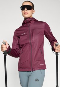 Maroon softshell jacket with hood, zippered pockets, and arm logo; paired with light blue pants featuring a logo. Black poles held in hands.