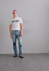Man står mot en grå vägg iförd en vit Moschino T-shirt, blå jeans med hjärtan och fyrkantiga lappar, samt svarta skor med vita snörningar.