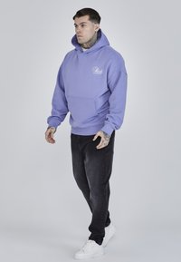 Sudadera de algodón morada con un bolsillo delantero y capucha con cordón, combinada con jeans negros y zapatillas blancas. Detalle gráfico sencillo en el pecho.
