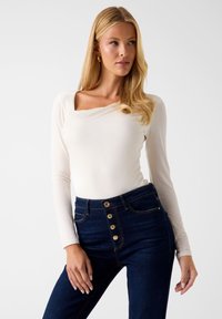 Langärmliges, cremeweißes Oberteil mit quadratischem Ausschnitt, kombiniert mit hoch taillierten, dunkelblauen Jeans mit goldfarbenen Knöpfen und glatter Textur.