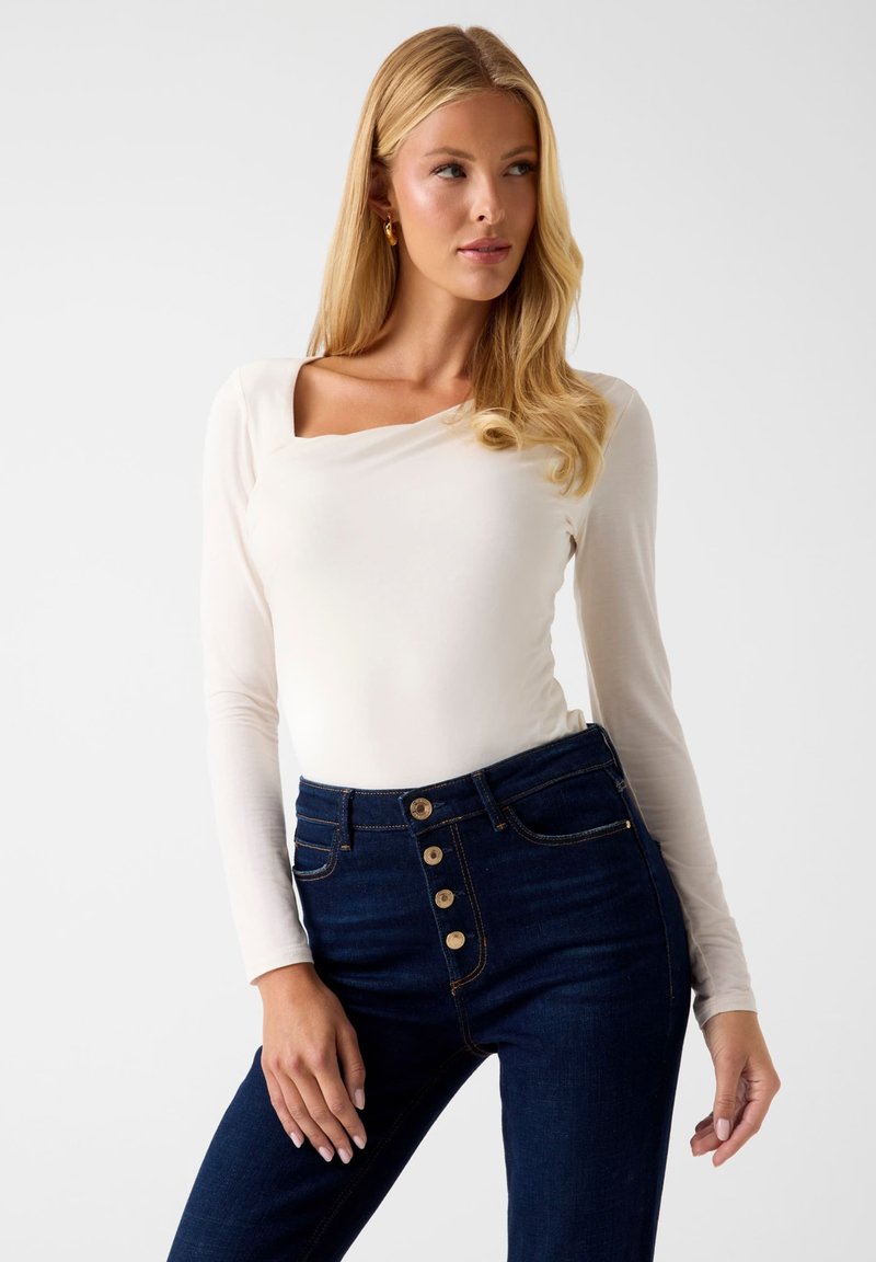 Langärmliges, cremeweißes Oberteil mit quadratischem Ausschnitt, kombiniert mit hoch taillierten, dunkelblauen Jeans mit goldfarbenen Knöpfen und glatter Textur.