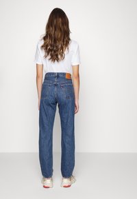 Blå jeans i hög midja med rak skärning, bakfickor och en brun läderlapp vid midjebandet. Texturerat tyg, minimalistisk design.