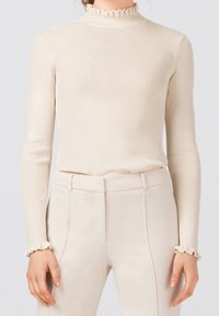 Kvinna som bär en krämfärgad ribbad långärmad topp med volanger vid den höga halsringningen och ärmarna, i kombination med högmidjade krämfärgade byxor.