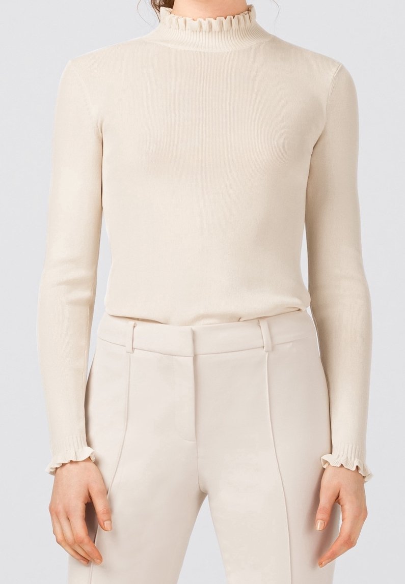 Kvinna som bär en krämfärgad ribbad långärmad topp med volanger vid den höga halsringningen och ärmarna, i kombination med högmidjade krämfärgade byxor.