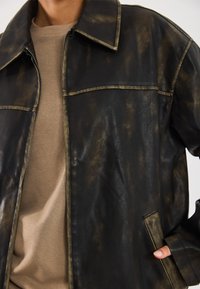 Gros plan sur une personne portant une veste en cuir marron foncé usée sur une chemise marron clair, avec une main dans la poche de la veste.