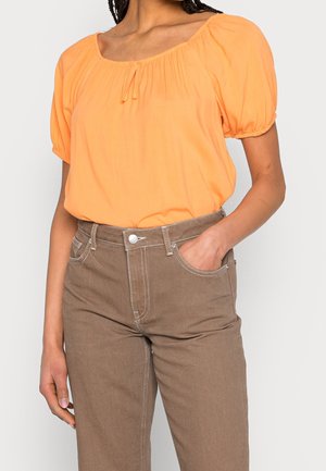 Kvinde iført en løs orange bluse med puffærmer stoppet ned i højtaljede brune bukser, med højre hånd i lommen.