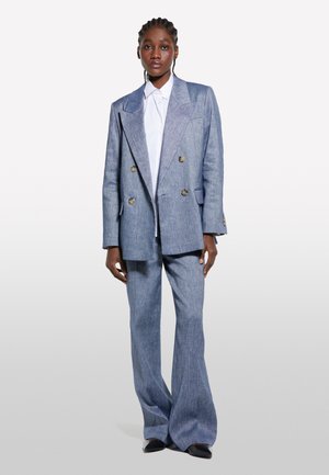 Un mannequin pose dans un costume bleu clair ajusté à double boutonnage avec un large pantalon et une chemise blanche, sur fond uni.
