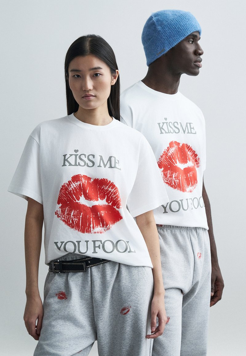 White t-shirts with red lipstick kiss print and text "KISS ME YOU FOOL," paired with gray sweatpants featuring similar kiss designs. Blue knitted beanie.