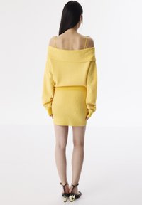 Pull en maille jaune dénudé sur les épaules associé à une mini-jupe assortie. Il présente un tissu texturé et des sandales noires à talons avec des accents en forme de cœur.